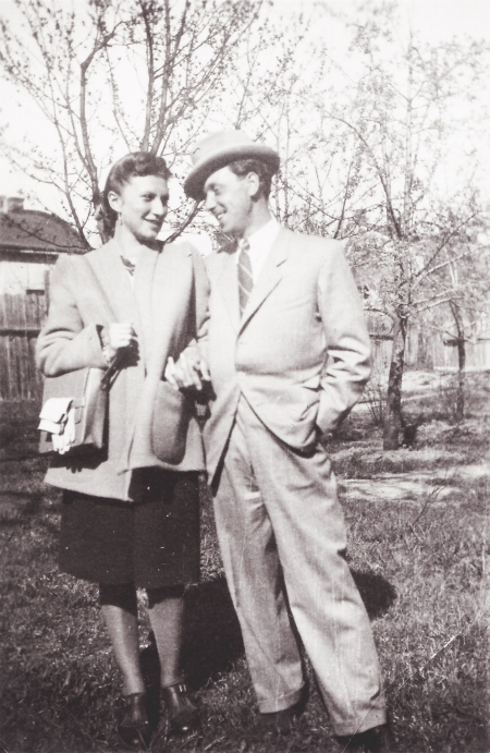 Photo en noir et blanc d'un homme et d'une femme, bras-dessus bras-dessous, sur un terrain extérieur avec des arbres en arrière-plan. Le couple se sourit l'un à l'autre et l'homme porte un complet et un chapeau.