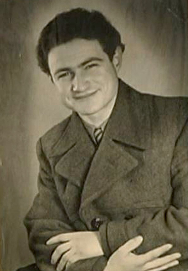 Black-and-white portrait photograph of a young man, pictured from the waist up, sitting with his arms crossed. He wears a jacket and smiles at the camera.