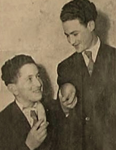 Photo en noir et blanc de deux jeunes hommes, l'un des deux est assis, qui se sourient l'un à l'autre. Les deux jeunes hommes portent des complets et tiennent ce qui semble être des morceaux de fruits.