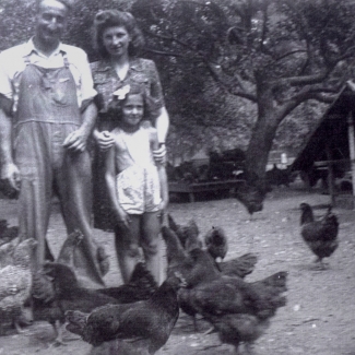Photo en noir et blanc d'un homme et d'une femme avec une jeune fille, se tenant parmi des douzaines de poulets dans une bassecour. Un poulaillier est visible en arrière-plan.