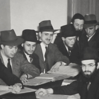 Black-and-white photograph of a group of 10 men sitting together at one table, covered in books and papers. The men wear hats and suits.