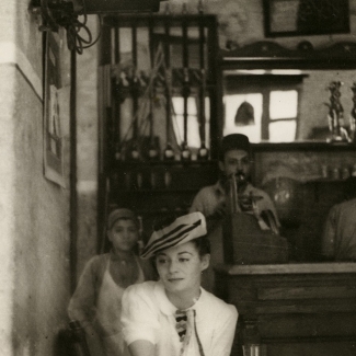 Photo en noir et blanc d'une femme assise à un café, portant une blouse blanche et un béret. La femme regarde de côté. un jeune garçon et deux hommes sont debout derrière le bar en arrière-plan.