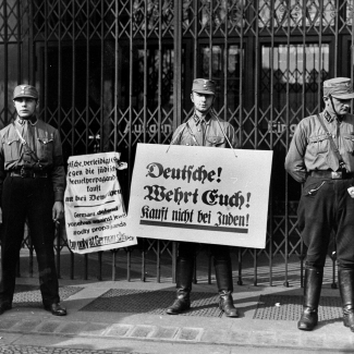 Photo en noir et blanc de trois hommes en uniforme militaire, se tenant devant une devanture de commerce fermée avec des portes en fer. Deux hommes tiennent des pancartes rédigées en allemand. L'autre soldat discute avec un civil.