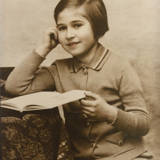 Sepia-tone photograph of a young girl, sitting at a desk with a tablecloth. The girl rests an open book on the desk with one hand, and leans on her other elbow in a pensive pose. Light pink shading has been added to her lips.