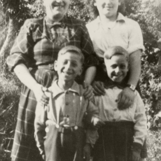 Black-and-white photograph of a two women standing behind two young twin boys, all 4 of them smiling at the camera. They are standing outside and in what looks like summer weather, due to the women’s rolled-up sleeves, and the boys’ shorts.
