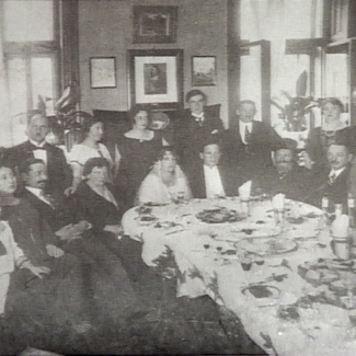 Photo en noir et blanc d'un groupe d'environ 20 personnes assises ensemble autour d'une grande table remplie de plats. Le groupe est habillé de manière formelle et un couple assis à la table semble célébré leur mariage.