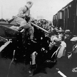 Black-and-white photograph of a man sitting on the edge of a train car, holding a cane, and looking onto a platform full of people.