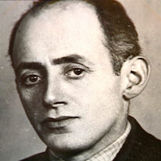 Portrait photographique du visage d'un homme qui regarde la caméra. Ses cheveux de couleur foncée sont coiffés vers l'arrière et il semble qu'il porte un complet.