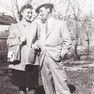 Photo en noir et blanc d'un homme et d'une femme, bras-dessus bras-dessous, sur un terrain extérieur avec des arbres en arrière-plan. Le couple se sourit l'un à l'autre et l'homme porte un complet et un chapeau.