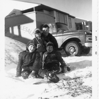 Photo en noir et blanc d'un homme avec trois jeunes enfants, assis à l'extérieur dans la neige. Ils sourient à la caméra, rassemblés ensemble devant une maison avec un camion en arrière-plan.