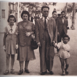Photo de couleur sépia d'un homme et d'une femme, tenant les mains de leurs deux enfants se tenant de chaque côté d'eux. La famille de quatre est à l'extérieur sur une rue avec des personnes qui marchent en arrière-plan.