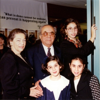 Photo en couleur d'un homme et d'une femme âgés se tenant ensemble à l'intérieur avec une autre femme et deux jeunes filles. La famille est habillée de manière formelle et les filles portent des blouses et l'homme un complet.
