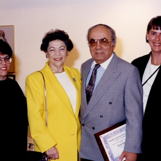 Photo en couleur d'un homme et d'une femme âgés se tenant entre deux autres femmes. Ils sourient tous les quatre à la caméra. L'homme porte un complet et tient un document encadré.
